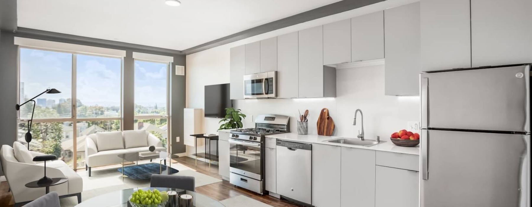 Bright apartment interior with a modern kitchen, stainless steel appliances, and a living area with large windows and white sofas.